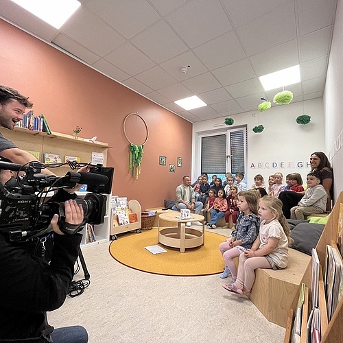 Eine Gruppe von rund 20 Kindern sitzt in einem Kita-Raum in mehreren Reihen auf Bänken und Kissen. Neben ihnen sitzen zwei Erwachsene. Im Vordergrund filmt ein Kamerateam die Szene mit einer großen Fernsehkamera.