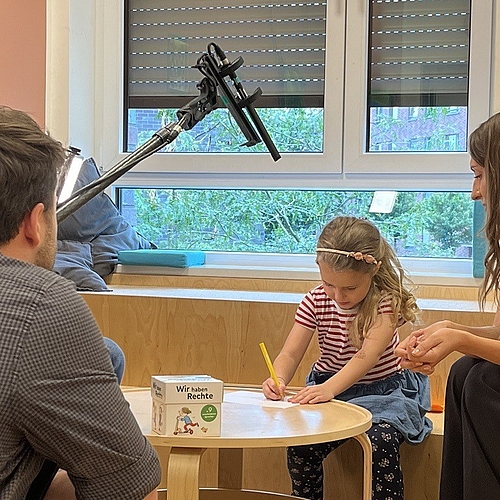 Ein Mädchen sitzt an einem kleinen Tisch in einer Kita und schreibt mit einem Bleistift auf ein Blatt Papier. Neben ihr sitzt eine Erzieherin und lächelt, während ein Kamerateam die Szene aufnimmt. Auf dem Tisch liegt ein Buch mit dem Titel „Wir haben Rechte“.